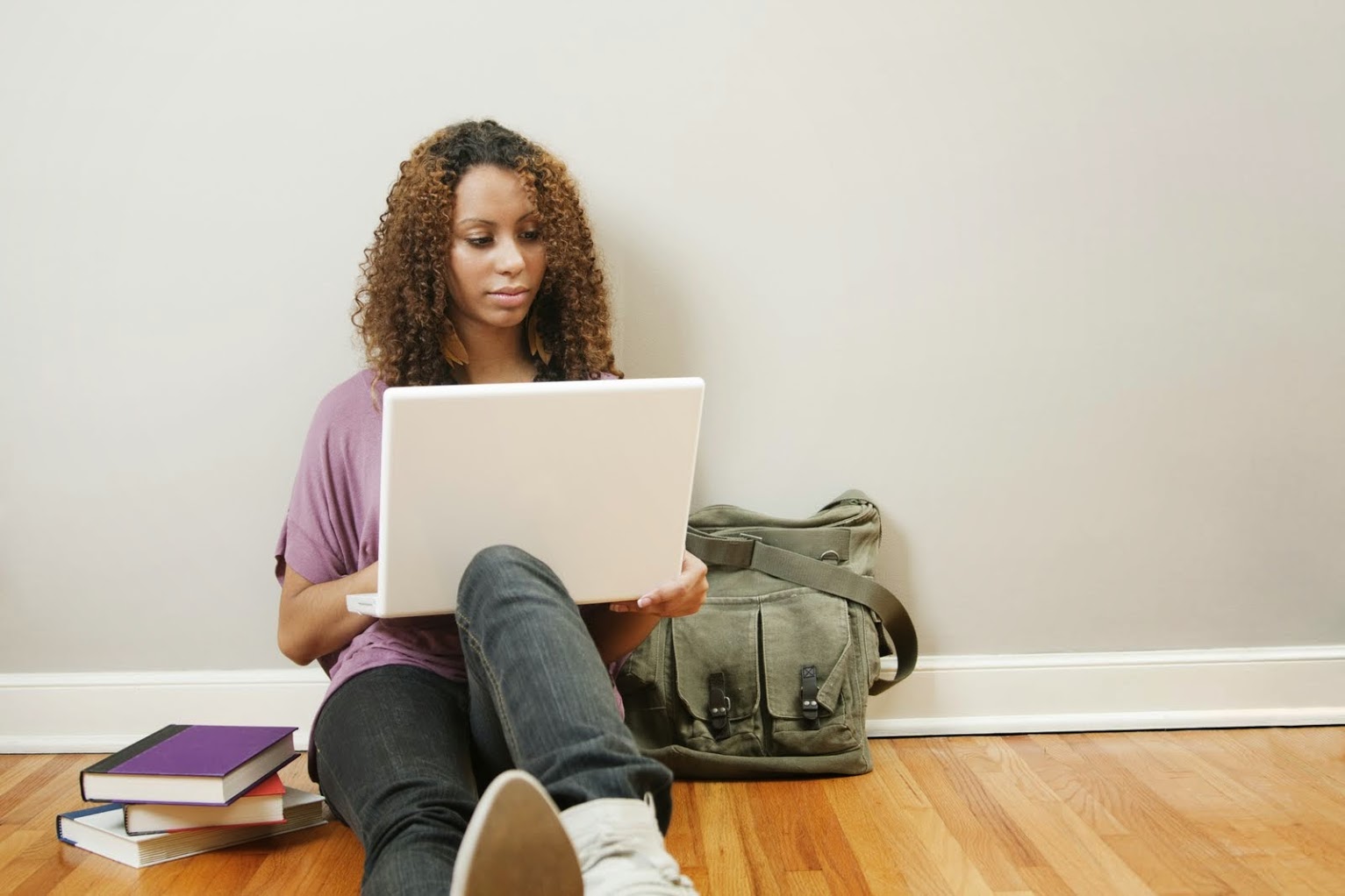 Chica sentada en el suelo de madera mientras se apoya contra una pared gris y mira su computadora portátil con un bolso verde junto a ella